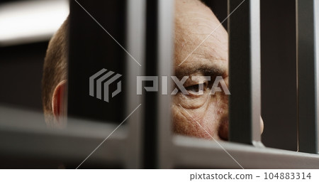 Face close up of depressed elderly criminal standing behind metal bars Face close up of depressed elderly criminal standing behind metal bars 104883314