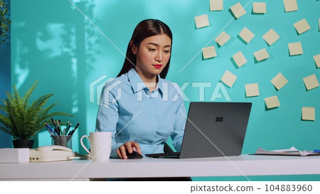 Smiling professional executive imputing data on laptop, drinking coffee at modern colourful office desk over blue studio background. Asian businesswoman satisfied with work results Smiling professional executive imputing data on laptop, drinking coffee at modern colourful office desk over blue studio background. Asian businesswoman satisfied with work results 104883960
