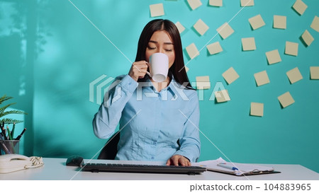 Asian businesswoman having cup of coffee while in online videoconference meeting, carefully listening and chatting using keyboard. Employee in modern office over blue studio background 104883965