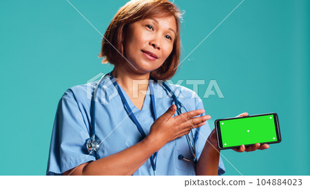 Close up shot of clinic employee isolated over studio background showing medical tape on chroma key phone green screen. Nurse holding smartphone in landscape mode, presenting informative video 104884023