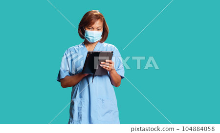 Lively asian nurse looking at patient data on digital tablet, confused with medical checkup results. Professional clinical worker wearing protective face mask, isolated over blue studio background Lively asian nurse looking at patient data on digital tablet, confused with medical checkup results. Professional clinical worker wearing protective face mask, isolated over blue studio background 104884058