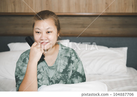 Asian housewife sit and smile in bed and holding remote control watching television with relaxing action. Hobby and free time life style concept. Asian housewife sit and smile in bed and holding remote control watching television with relaxing action. Hobby and free time life style concept. 104887706