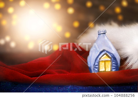 Decorative ice house on Santa Claus's red velvet hat with fur. Blurred lights with lens flare. Christmas, New Year postcard. Copy spac Decorative ice house on Santa Claus's red velvet hat with fur. Blurred lights with lens flare. Christmas, New Year postcard. Copy spac 104888256