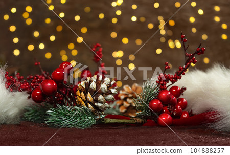 Spruce branches with red berries and pine cones on a red Santa Claus' hat with new year lights in the back. Blurred background. Christmas card 104888257