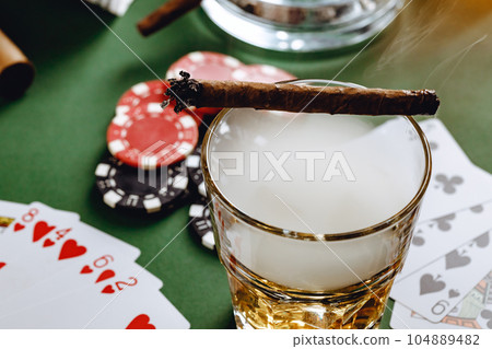 Glass of whiskey, cigar, playing cards and chips on green background Glass of whiskey, cigar, playing cards and chips on green background 104889482