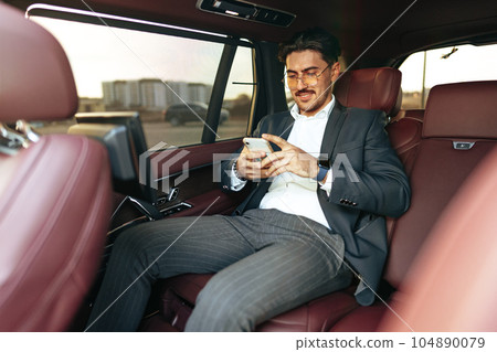 Young elegant businessman sitting on the back seat of a luxury car and using smartphone 104890079