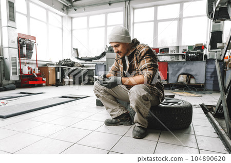 Car service worker sitting on a wheel and resting, using mobile phone 104890620
