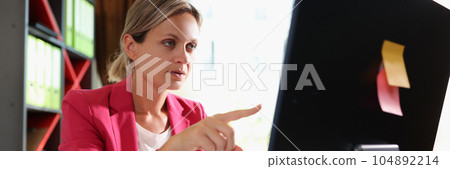 Business woman with serious face at office desk points her finger at computer screen. Business woman with serious face at office desk points her finger at computer screen. 104892214