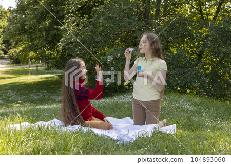 Joyful Asian Caucasian Girls, Children Of Elementary School Age Blow Soap Bubbles In Summer Park, Meadow. Happy Childhood. School Break Time. Children Protection Day, Earth Day. Horizontal Plane Joyful Asian Caucasian Girls, Children Of Elementary School Age Blow Soap Bubbles In Summer Park, Meadow. Happy Childhood. School Break Time. Children Protection Day, Earth Day. Horizontal Plane 104893060