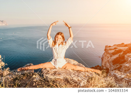 Happy smiling teen girl balerina performs stretching exercises splits on the beach during a beautiful sunset, enjoying a sunny summer day. Happy smiling teen girl balerina performs stretching exercises splits on the beach during a beautiful sunset, enjoying a sunny summer day. 104893334