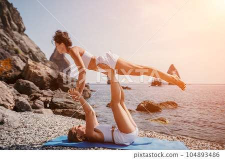 Woman sea yoga. Two happy women practicing yoga on the beach with ocean and rock mountains. Motivation and inspirational fit and exercising. Healthy lifestyle outdoors in nature, fitness concept. 104893486