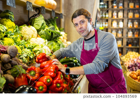 Male grocery store worker arranges bell peppers and other vegetables on the counter and display case 104893569