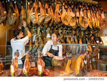 Positive young man and woman selling spanish jamon at counter Positive young man and woman selling spanish jamon at counter 104893591