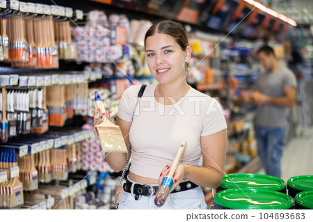 Positive female customer chooses paint brushes on the shelves of hardware store Positive female customer chooses paint brushes on the shelves of hardware store 104893683