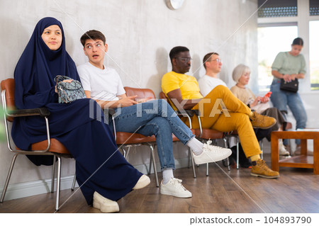 Man communicates with an Asian woman in a veil while waiting for a specialist in the office along with other people of nationality 104893790