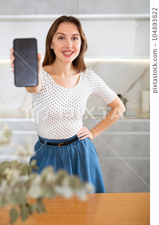 Middle-aged woman showing her smartphone standing in the kitchen 104893822