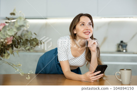 Young housewife leaned on countertop, and with cup of coffee rests and sticks in phone Young housewife leaned on countertop, and with cup of coffee rests and sticks in phone 104894150