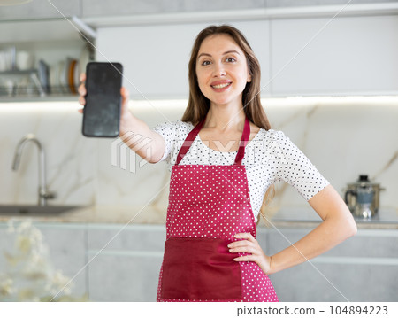 Middle-aged woman wearing apron showing her smartphone standing in the kitchen Middle-aged woman wearing apron showing her smartphone standing in the kitchen 104894223