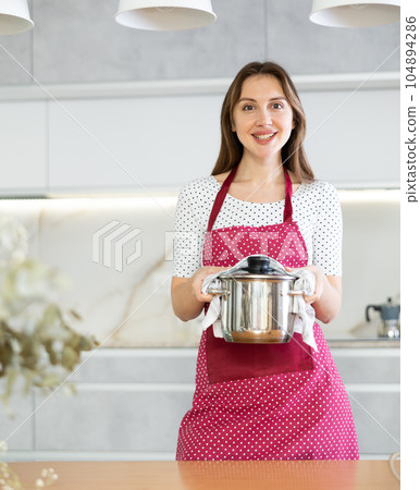 Smiling girl is standing in kitchen in dotted apron and holding stainless steel pot with lid 104894286