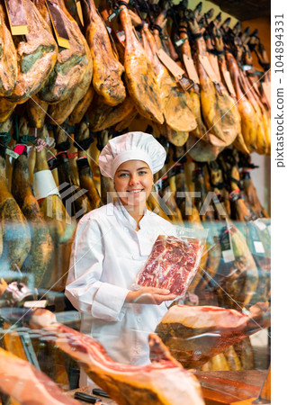 Female seller cutting slices from whole leg of jamon in meat store 104894331