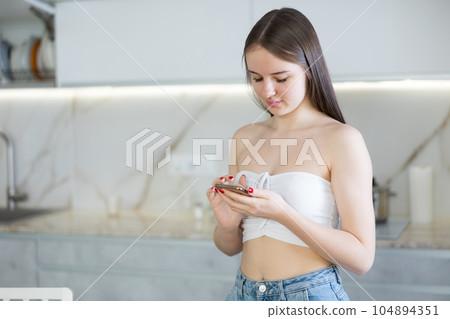 Bored young woman stands in kitchen leans on countertop and surfs internet. Home pastime 104894351