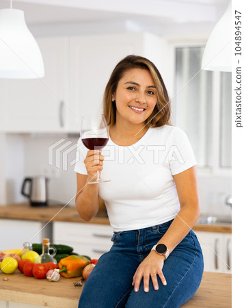 Smiling young Hispanic woman drinking wine sitting on table in kitchen 104894547