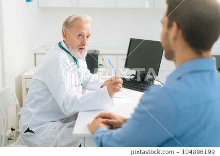 Senior adult male doctor talking with unrecognizable male patient during medical visit sitting at table in medical office room. Mature older physician consulting man at hospital. 104896199