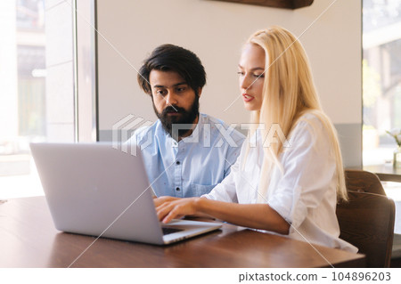 Medium shot of two business partners discussing actively startup project sitting in cozy cafe by window. Bearded businessman explaining project to female colleague using at laptop computer. 104896203