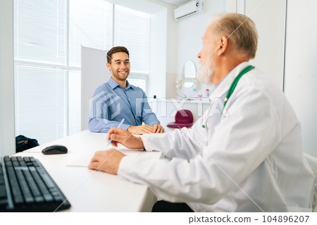 Cheerful male patient talking with mature adult doctor sitting at table during checkup visit in medical clinic office. Happy young man client listening to older senior physician explaining treatment. Cheerful male patient talking with mature adult doctor sitting at table during checkup visit in medical clinic office. Happy young man client listening to older senior physician explaining treatment. 104896207