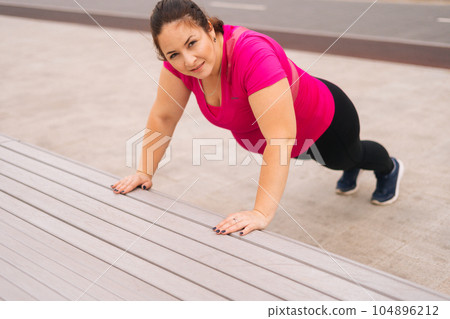 Top view of positive fat young woman training doing push ups on street bench in city park at summer morning, looking at camera. Fitness female doing bench push ups for strength workout outdoors. Top view of positive fat young woman training doing push ups on street bench in city park at summer morning, looking at camera. Fitness female doing bench push ups for strength workout outdoors. 104896212