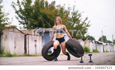 sexy athletic young blond woman in shorts, performs various strength exercises with the help of tires, lifting weights. In summer, near old abandoned garages sexy athletic young blond woman in shorts, performs various strength exercises with the help of tires, lifting weights. In summer, near old abandoned garages 104897525