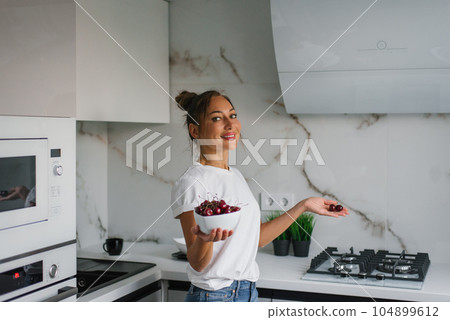 Pretty smiling young woman holds a plate of fresh cherry berries in her hand in the kitchen 104899612