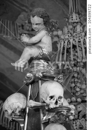 Grayscale shot of an angel sculpture on a stack of skulls in the Sedlec Ossuary in Kutna Hora Grayscale shot of an angel sculpture on a stack of skulls in the Sedlec Ossuary in Kutna Hora 104899722