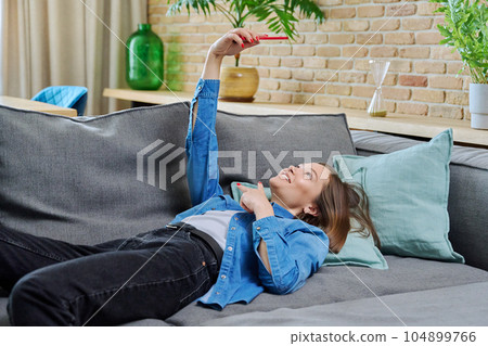 Young woman having video call lying on sofa at home, using smartphone Young woman having video call lying on sofa at home, using smartphone 104899766