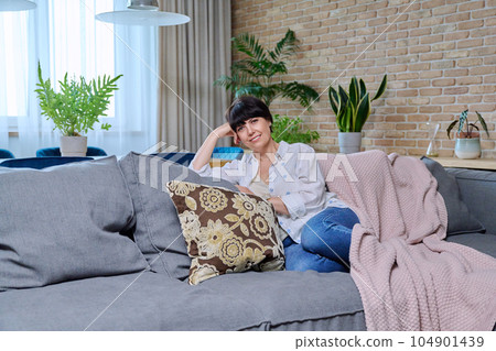 Beautiful smiling mature woman sitting on sofa in living room at home Beautiful smiling mature woman sitting on sofa in living room at home 104901439