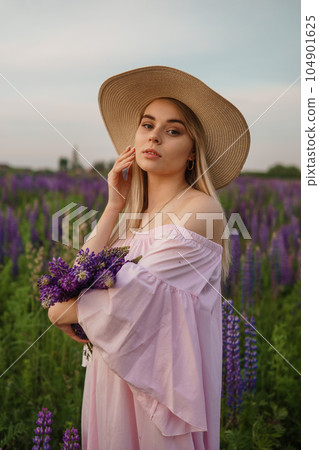 A beautiful woman in a straw hat walks in a field with purple flowers. A walk in nature in the lupin field A beautiful woman in a straw hat walks in a field with purple flowers. A walk in nature in the lupin field 104901625