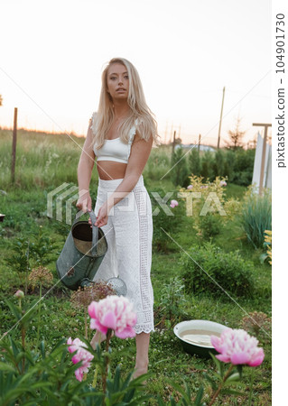 A blonde woman is watering flowers in the garden in the courtyard of a village house. A blonde woman is watering flowers in the garden in the courtyard of a village house. 104901730