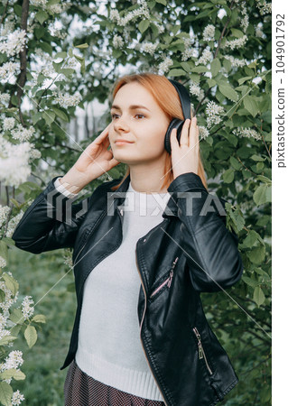 A red-haired teenage girl listens to music in headphones, configured via the phone. The time of spring flowering of cherry orchards A red-haired teenage girl listens to music in headphones, configured via the phone. The time of spring flowering of cherry orchards 104901792