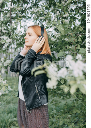 A red-haired teenage girl listens to music in headphones, configured via the phone. The time of spring flowering of cherry orchards 104901793