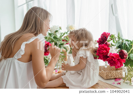 A little blonde girl with her mom on a kitchen countertop decorated with peonies. The concept of the relationship between mother and daughter. Spring atmosphere. A little blonde girl with her mom on a kitchen countertop decorated with peonies. The concept of the relationship between mother and daughter. Spring atmosphere. 104902041