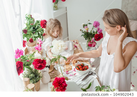 A little blonde girl with her mom on a kitchen countertop decorated with peonies. The concept of the relationship between mother and daughter. Spring atmosphere. A little blonde girl with her mom on a kitchen countertop decorated with peonies. The concept of the relationship between mother and daughter. Spring atmosphere. 104902043