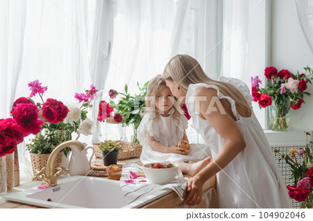 A little blonde girl with her mom on a kitchen countertop decorated with peonies. The concept of the relationship between mother and daughter. Spring atmosphere. A little blonde girl with her mom on a kitchen countertop decorated with peonies. The concept of the relationship between mother and daughter. Spring atmosphere. 104902046