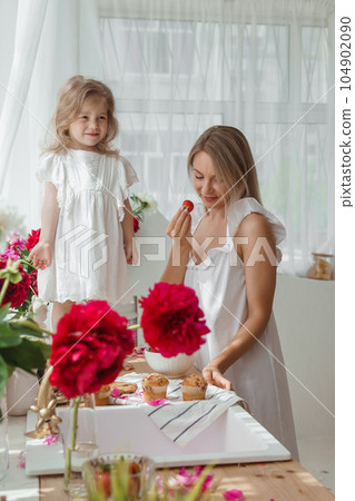 A little blonde girl with her mom on a kitchen countertop decorated with peonies. The concept of the relationship between mother and daughter. Spring atmosphere. A little blonde girl with her mom on a kitchen countertop decorated with peonies. The concept of the relationship between mother and daughter. Spring atmosphere. 104902090