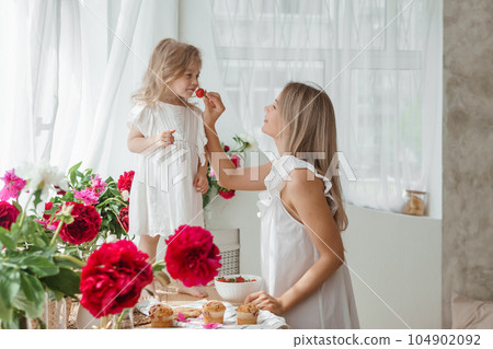 A little blonde girl with her mom on a kitchen countertop decorated with peonies. The concept of the relationship between mother and daughter. Spring atmosphere. A little blonde girl with her mom on a kitchen countertop decorated with peonies. The concept of the relationship between mother and daughter. Spring atmosphere. 104902092