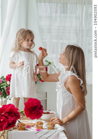 A little blonde girl with her mom on a kitchen countertop decorated with peonies. The concept of the relationship between mother and daughter. Spring atmosphere. A little blonde girl with her mom on a kitchen countertop decorated with peonies. The concept of the relationship between mother and daughter. Spring atmosphere. 104902093