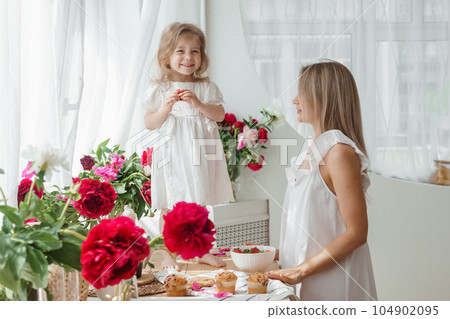 A little blonde girl with her mom on a kitchen countertop decorated with peonies. The concept of the relationship between mother and daughter. Spring atmosphere. A little blonde girl with her mom on a kitchen countertop decorated with peonies. The concept of the relationship between mother and daughter. Spring atmosphere. 104902095