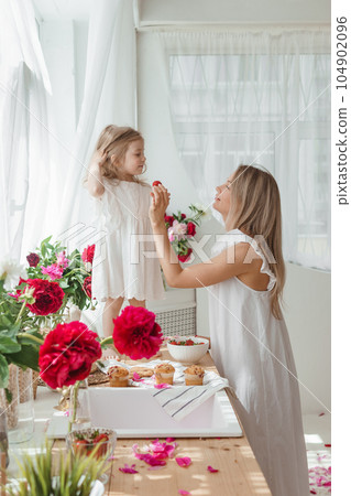A little blonde girl with her mom on a kitchen countertop decorated with peonies. The concept of the relationship between mother and daughter. Spring atmosphere. A little blonde girl with her mom on a kitchen countertop decorated with peonies. The concept of the relationship between mother and daughter. Spring atmosphere. 104902096