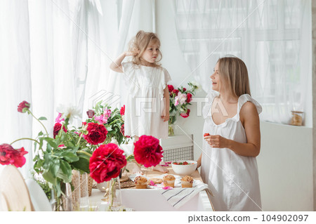 A little blonde girl with her mom on a kitchen countertop decorated with peonies. The concept of the relationship between mother and daughter. Spring atmosphere. A little blonde girl with her mom on a kitchen countertop decorated with peonies. The concept of the relationship between mother and daughter. Spring atmosphere. 104902097