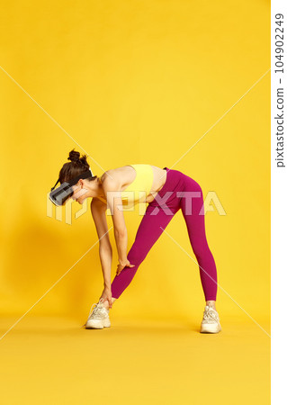 Full-length image of young slim woman wearing vr glases and training against yellow studio background. Warming-up, stretching 104902249