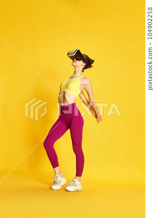 Full-length image of young sportive woman wearing vr glases and training against yellow studio background. Warming-up, stretching 104902258
